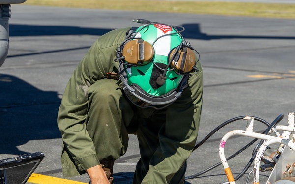 A military aviation technician in olive green flight suit and green helmet is crouched on a tarmac performing maintenance work beneath an aircraft. The technician is working with hydraulic or pneumatic fittings connected to a pressurized cylinder mounted in a white metal cage frame. A ruggedized military laptop is open on the ground nearby, likely used for diagnostics or technical reference.