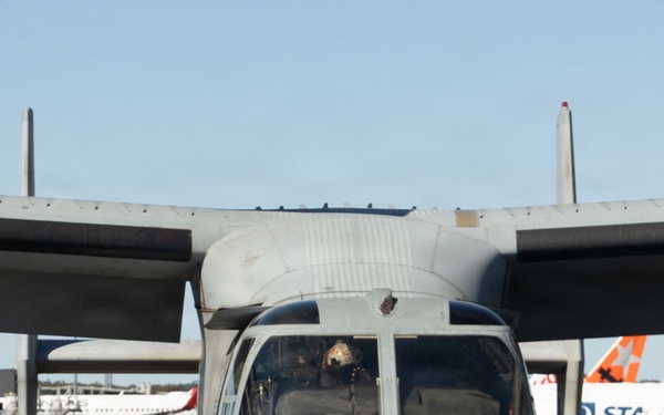 A military tiltrotor aircraft, identifiable as a Bell Boeing V-22 Osprey, is parked on a civilian airport tarmac. A flight crew member in olive green flight suit and white helmet stands at the nose of the aircraft, apparently conducting a pre-flight or post-flight inspection. In the background, Qantas commercial aircraft are visible, indicating a shared civilian-military airport environment.