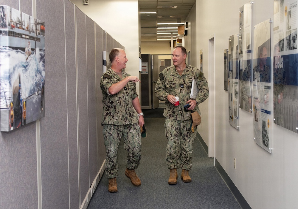 Two U.S. Navy personnel in NWU Type I (digital blue/green camouflage) uniforms are conversing in an office corridor. One individual is pointing toward photographs displayed on a partition wall while the other listens and smiles. The hallway features framed informational displays and photographs mounted on the right wall, suggesting a command or administrative facility.