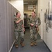 Two U.S. Navy personnel in NWU Type I (digital blue/green camouflage) uniforms are conversing in an office corridor. One individual is pointing toward photographs displayed on a partition wall while the other listens and smiles. The hallway features framed informational displays and photographs mounted on the right wall, suggesting a command or administrative facility.