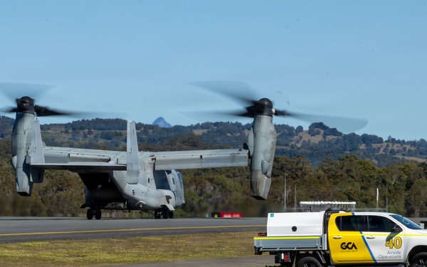 A military tiltrotor aircraft, identifiable as a Bell Boeing V-22 Osprey, is captured mid-hover or landing on an airfield with rotors in motion. In the foreground, a white and yellow utility pickup truck marked 'GCA Airside Operations' with the number '40' is parked on the tarmac. The setting appears to be a civilian or joint-use airport surrounded by green hills and trees under clear blue skies.