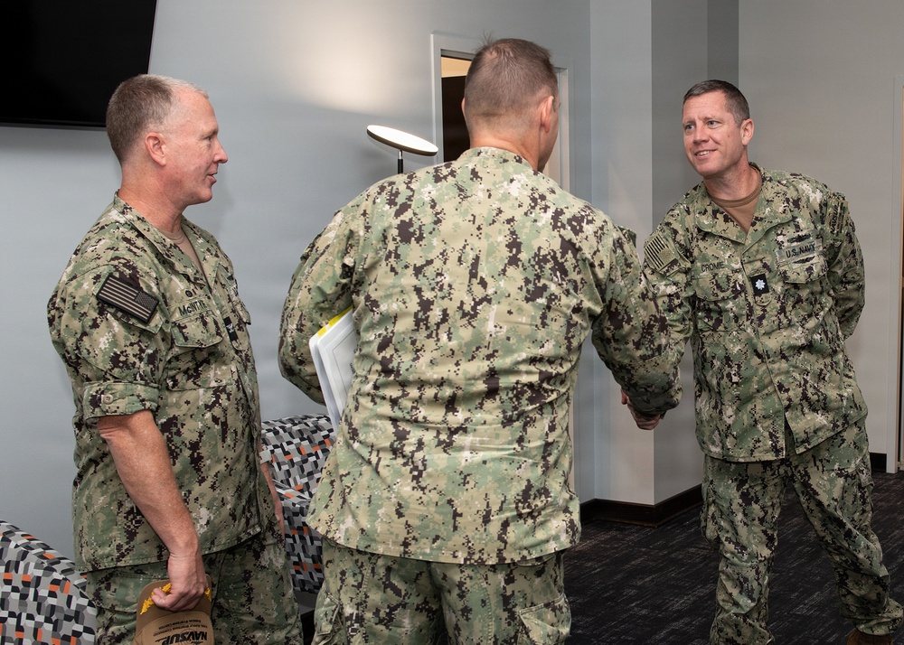 Three U.S. Navy personnel in NWU Type III (green digital camouflage) uniforms are engaged in a meeting or greeting interaction indoors. Two individuals are facing the camera while one has his back turned, appearing to shake hands with the person on the right. The setting appears to be a modern office or conference room environment.