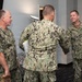 Three U.S. Navy personnel in NWU Type III (green digital camouflage) uniforms are engaged in a meeting or greeting interaction indoors. Two individuals are facing the camera while one has his back turned, appearing to shake hands with the person on the right. The setting appears to be a modern office or conference room environment.