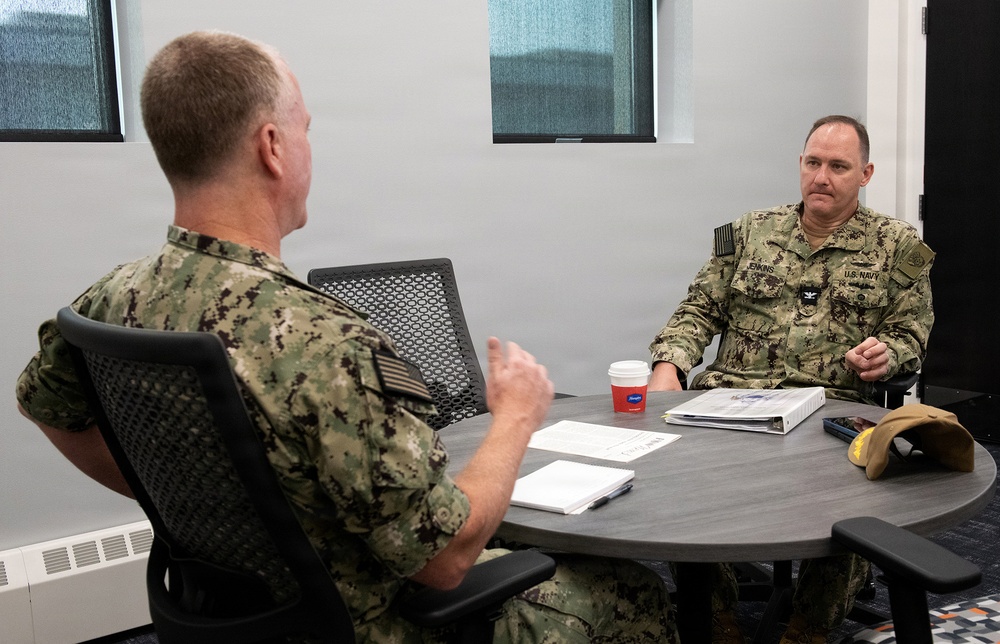 Two U.S. Navy personnel in NWU Type III (woodland digital camouflage) uniforms are seated at a round conference table engaged in a discussion. The individual facing the camera has a name tape reading 'JENKINS' and 'U.S. NAVY' with what appears to be a Captain's eagle insignia. The setting appears to be a modern office or conference room environment.