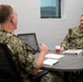 Two U.S. Navy personnel in NWU Type III (woodland digital camouflage) uniforms are seated at a round conference table engaged in a discussion. The individual facing the camera has a name tape reading 'JENKINS' and 'U.S. NAVY' with what appears to be a Captain's eagle insignia. The setting appears to be a modern office or conference room environment.