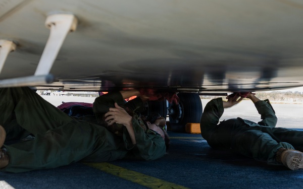 Two individuals in military-style green flight suits are lying on an airfield tarmac beneath the fuselage of an aircraft. They appear to be conducting an inspection or maintenance check of the aircraft's underside. The image is taken from ground level, providing a low-angle perspective of the scene.
