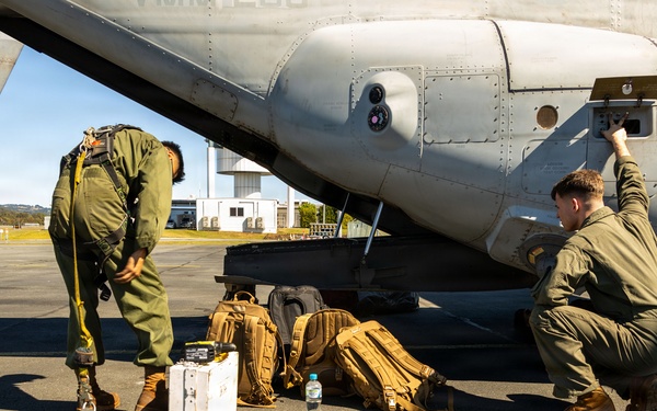 Military personnel in olive/green flight suits are conducting pre- or post-flight activities on an airport tarmac beside a large military aircraft. Three individuals are visible performing various tasks including equipment checks, gear management, and accessing an external panel on the aircraft fuselage. Multiple tactical backpacks, a white equipment case, and a flight helmet are staged on the ground nearby.