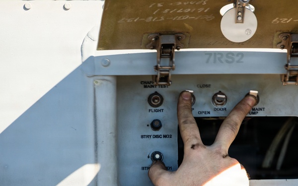 A person's hand is interacting with an external control panel on what appears to be a military or aerospace vehicle. The panel, labeled '7RS2', contains multiple switches and controls including battery disconnect switches and door controls. The panel is mounted on a light gray metal surface with riveted panels and a circular ventilation grille nearby.