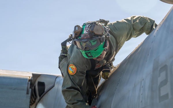 A military aviation technician or crew member in olive green flight suit is performing a pre-flight or maintenance inspection on a military aircraft. The individual is positioned on the fuselage of the aircraft, leaning forward to inspect or service a specific area. The setting appears to be an outdoor flight line or deck environment under clear skies.