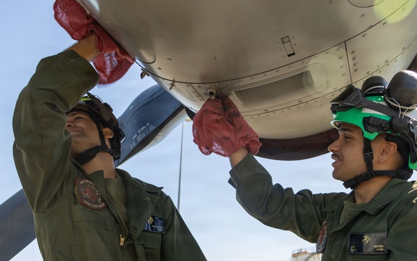 Two military personnel in olive green flight suits are performing maintenance or pre-flight inspection on an aircraft engine/propeller assembly on an airfield tarmac. Both individuals are using red shop rags to clean or inspect the engine nacelle area. One person wears a green helmet with ear protection, while the other wears a black helmet.