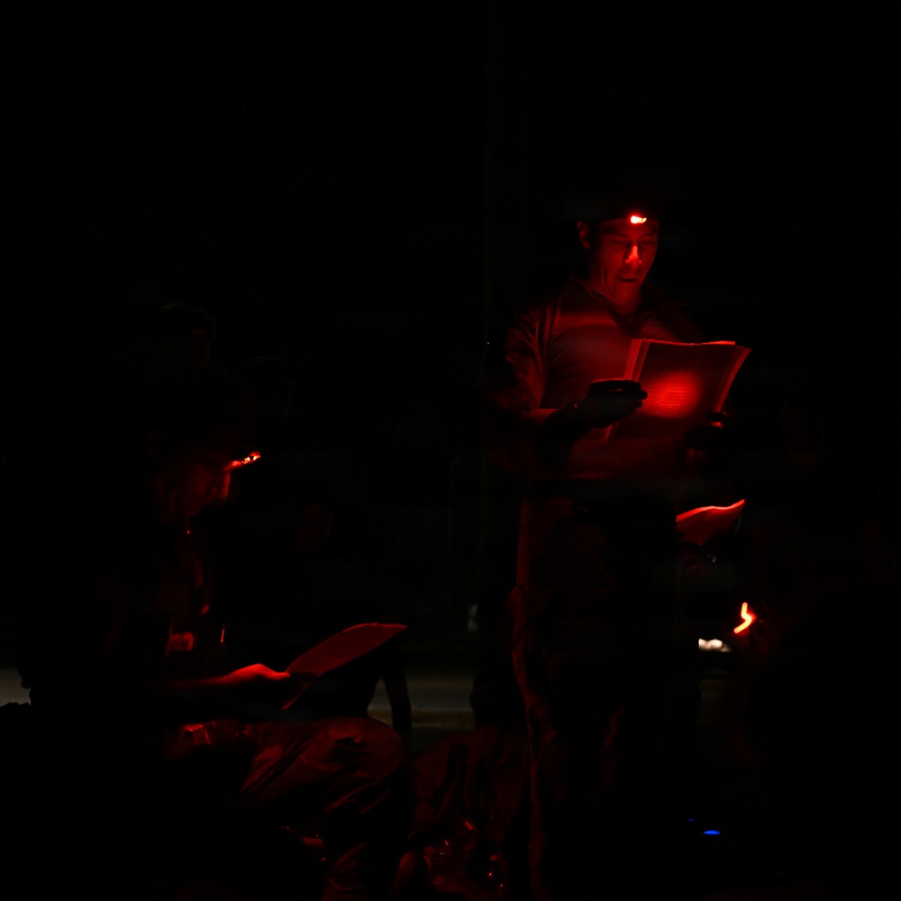 A very dark nighttime scene illuminated primarily by red light sources. At least two individuals are visible, with one standing person appearing to read or review documents using a red headlamp. The use of red lighting suggests a context where night vision preservation is important, such as military, astronomical, or field operations.
