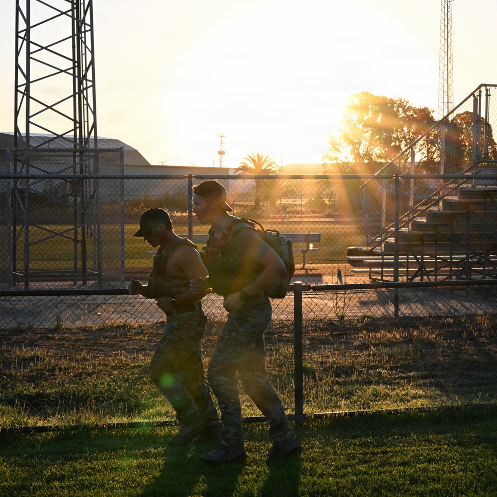 Two shirtless military personnel in camouflage pants and caps are conducting a ruck march or physical training exercise at what appears to be a military installation during golden hour. One individual is carrying a military-style rucksack on their back. The backlit sunset creates a strong silhouette effect against the facility's infrastructure.