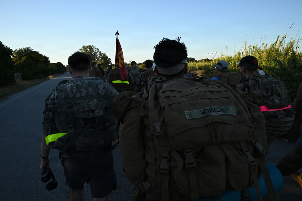 A group of military personnel or ROTC cadets are conducting a road march along a paved road at dawn or dusk. The group is moving away from the camera, carrying heavy rucksacks with name tapes visible, and a unit guidon (red/yellow flag) is being carried at the front of the formation. High-visibility reflective bands are worn by some participants, suggesting a safety-conscious organized march.