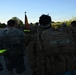 A group of military personnel or ROTC cadets are conducting a road march along a paved road at dawn or dusk. The group is moving away from the camera, carrying heavy rucksacks with name tapes visible, and a unit guidon (red/yellow flag) is being carried at the front of the formation. High-visibility reflective bands are worn by some participants, suggesting a safety-conscious organized march.