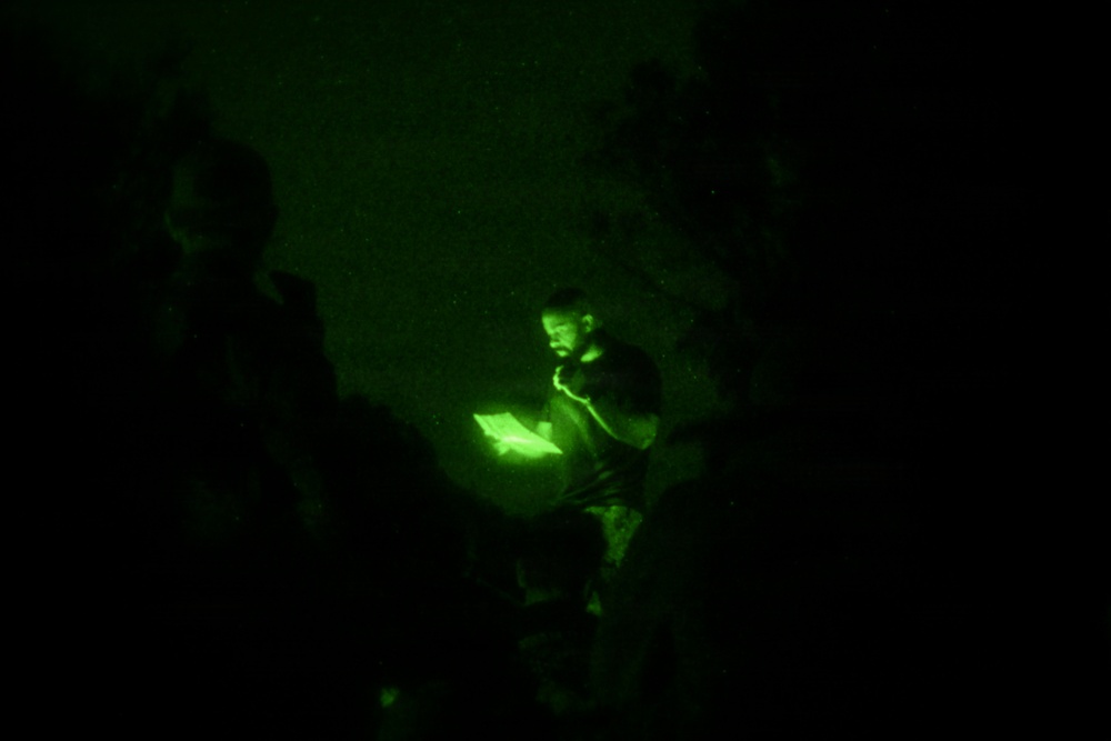 A night vision photograph showing a person illuminated by a bright light source, likely a tablet or document, in an otherwise dark environment. The green monochromatic tones are characteristic of night vision optics or infrared photography. Several silhouetted figures are visible in the foreground and background.