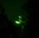A night vision photograph showing a person illuminated by a bright light source, likely a tablet or document, in an otherwise dark environment. The green monochromatic tones are characteristic of night vision optics or infrared photography. Several silhouetted figures are visible in the foreground and background.