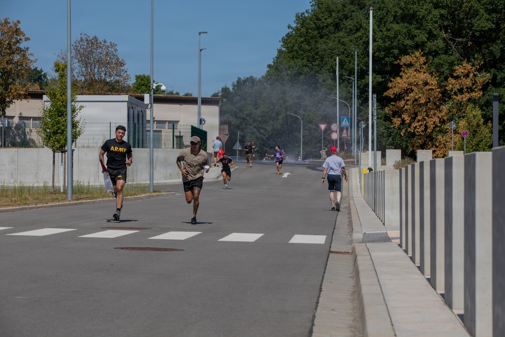 A group of people are running along a road on what appears to be a military installation or base, suggested by the 'ARMY' text visible on the lead runner's shirt. The scene shows multiple runners at various distances along the road, with smoke visible in the background, possibly from a smoke canister used in a training exercise or race event. The setting includes security walls, street lighting, road signs, and mature trees under a clear blue sky.