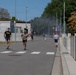 A group of people are running along a road on what appears to be a military installation or base, suggested by the 'ARMY' text visible on the lead runner's shirt. The scene shows multiple runners at various distances along the road, with smoke visible in the background, possibly from a smoke canister used in a training exercise or race event. The setting includes security walls, street lighting, road signs, and mature trees under a clear blue sky.