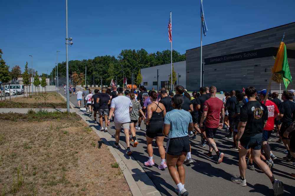 A large group of people in athletic wear are participating in a run or walk event near Sembach Elementary School, likely on a U.S. military installation in Germany. The crowd is moving along a sidewalk and road in warm, sunny weather. Flags, including an American flag and what appears to be unit or organizational flags, are visible along the route.