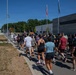 A large group of people in athletic wear are participating in a run or walk event near Sembach Elementary School, likely on a U.S. military installation in Germany. The crowd is moving along a sidewalk and road in warm, sunny weather. Flags, including an American flag and what appears to be unit or organizational flags, are visible along the route.