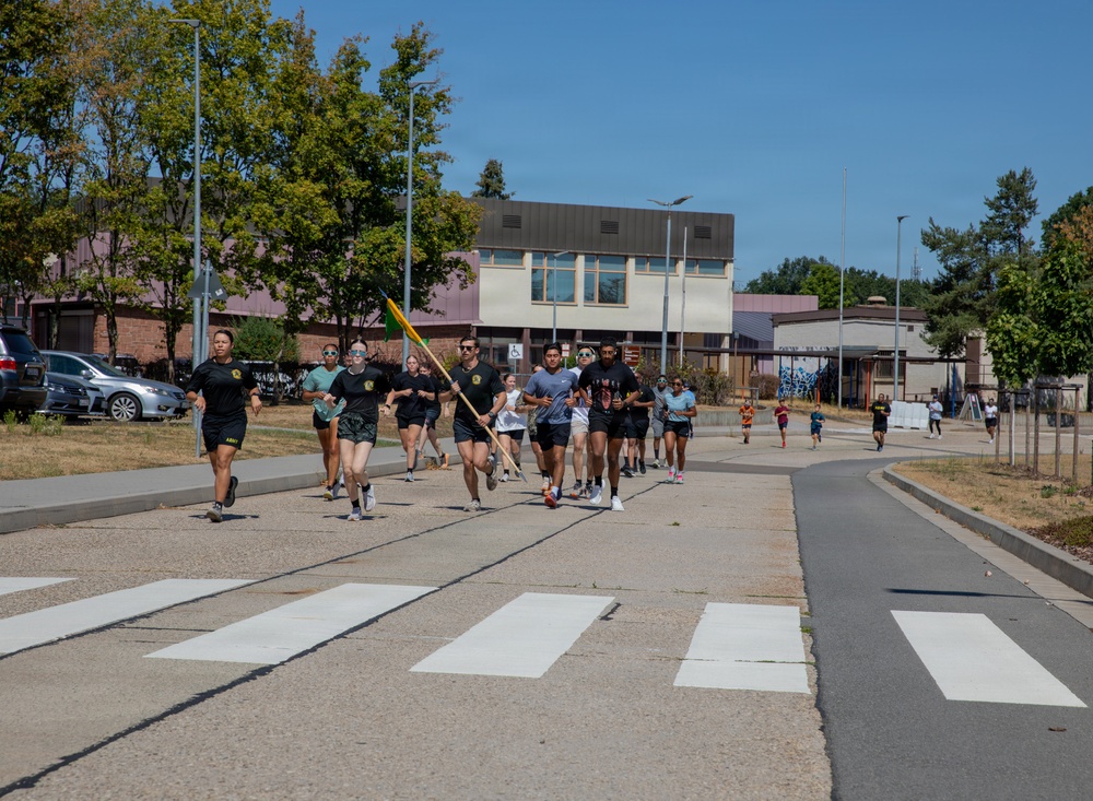 A group of runners, appearing to be military personnel based on their black PT (physical training) uniforms with unit patches, are running together in a formation across a crosswalk on what appears to be a military installation. One runner in the center is carrying a yellow-green guidon flag, which is characteristic of military unit runs. The event takes place on a sunny day with institutional buildings and trees visible in the background.