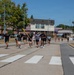 A group of runners, appearing to be military personnel based on their black PT (physical training) uniforms with unit patches, are running together in a formation across a crosswalk on what appears to be a military installation. One runner in the center is carrying a yellow-green guidon flag, which is characteristic of military unit runs. The event takes place on a sunny day with institutional buildings and trees visible in the background.