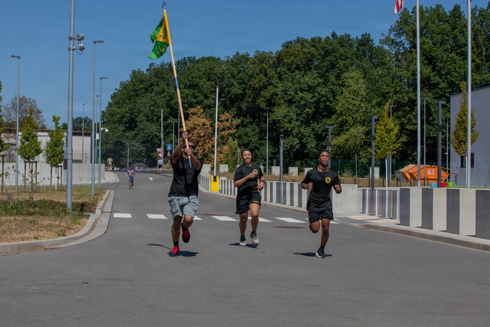 Three individuals are running along a paved road in what appears to be a military installation or base, with the lead runner carrying a green and yellow unit guidon flag. The runners are dressed in black athletic wear with military-style insignia visible on their shirts. The setting features a well-maintained facility with flagpoles, street lights, trees, and security barriers in the background.