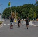 Three individuals are running along a paved road in what appears to be a military installation or base, with the lead runner carrying a green and yellow unit guidon flag. The runners are dressed in black athletic wear with military-style insignia visible on their shirts. The setting features a well-maintained facility with flagpoles, street lights, trees, and security barriers in the background.