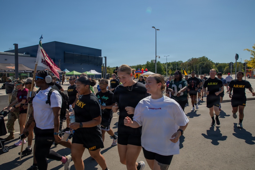 A group of U.S. Army personnel and civilians are participating in an outdoor run or fun run event on a sunny day. Several participants are wearing black Army PT (Physical Training) uniforms with 'ARMY' printed in gold letters, while others wear civilian clothing. The event appears to be a community or military fitness event with tents, inflatables, and spectators visible in the background.