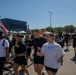 A group of U.S. Army personnel and civilians are participating in an outdoor run or fun run event on a sunny day. Several participants are wearing black Army PT (Physical Training) uniforms with 'ARMY' printed in gold letters, while others wear civilian clothing. The event appears to be a community or military fitness event with tents, inflatables, and spectators visible in the background.