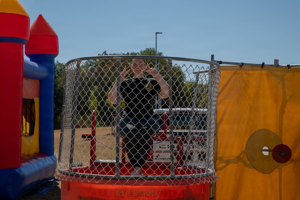 A person wearing a black shirt is seated inside a dunk tank, smiling and making peace signs with both hands. The dunk tank, branded 'Easy Dunker,' is surrounded by chain-link mesh and features a target mechanism on the right side. The scene appears to be an outdoor carnival or community event, with a colorful inflatable bounce house visible to the left.