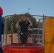 A person wearing a black shirt is seated inside a dunk tank, smiling and making peace signs with both hands. The dunk tank, branded 'Easy Dunker,' is surrounded by chain-link mesh and features a target mechanism on the right side. The scene appears to be an outdoor carnival or community event, with a colorful inflatable bounce house visible to the left.