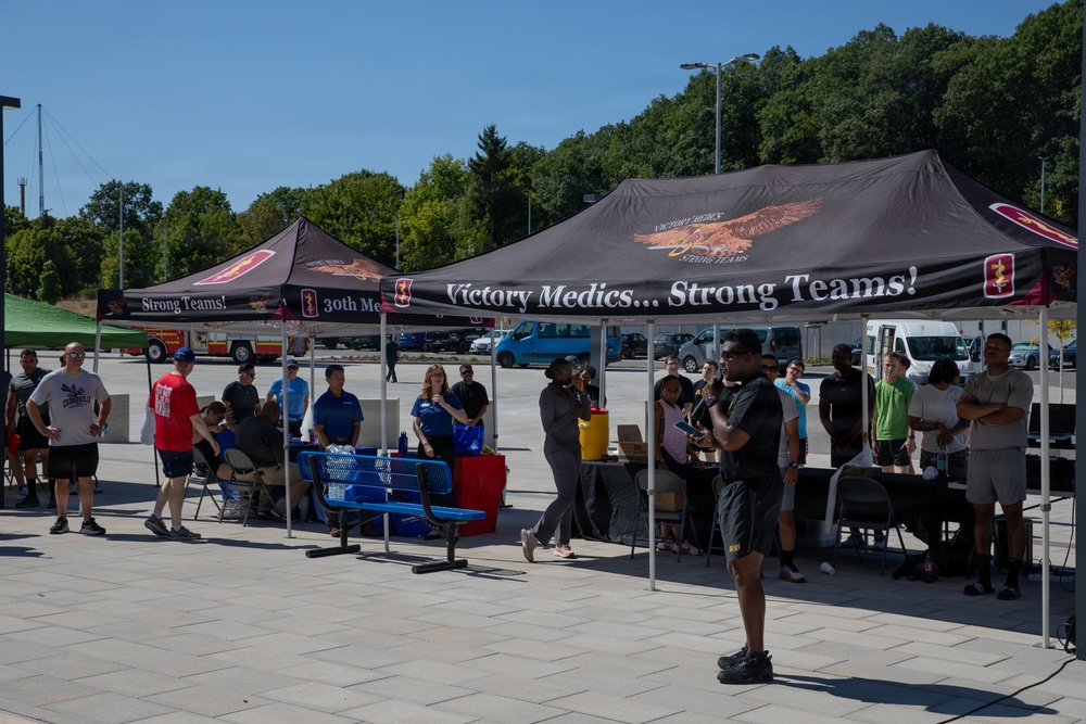 An outdoor event is taking place in a paved area, organized under two dark canopy tents bearing the slogan 'Victory Medics... Strong Teams!' and what appears to be '30th Me[dical]' branding. A person in dark clothing appears to be addressing or briefing a group of approximately 20 people gathered beneath and around the tents. The setting appears to be a military installation or large facility parking area, with trees and vehicles visible in the background.
