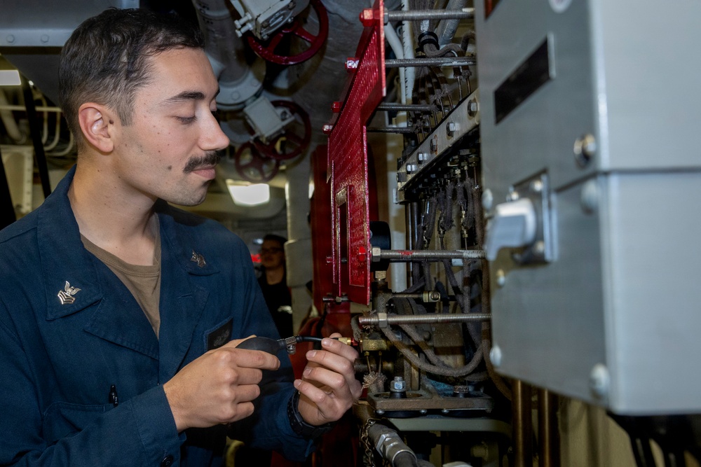 A young male U.S. Navy sailor in dark blue coveralls is performing maintenance or repair work on mechanical/piping equipment aboard a naval vessel. He is using a small tool, likely a screwdriver or wrench, to work on a fitting or valve assembly. The setting appears to be an engineering or machinery space below decks on a ship.