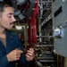 A young male U.S. Navy sailor in dark blue coveralls is performing maintenance or repair work on mechanical/piping equipment aboard a naval vessel. He is using a small tool, likely a screwdriver or wrench, to work on a fitting or valve assembly. The setting appears to be an engineering or machinery space below decks on a ship.