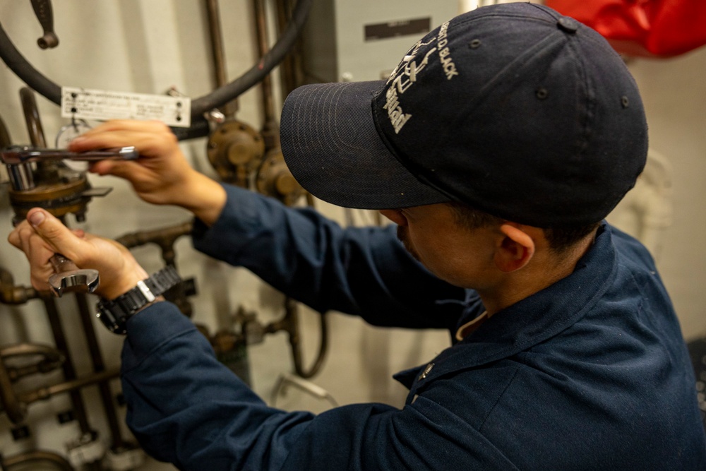 A technician wearing a navy blue uniform and baseball cap is performing maintenance work on a complex pipe and valve assembly. The individual is using two tools simultaneously — a wrench in one hand and what appears to be a screwdriver or pen-style tool in the other. The setting appears to be an industrial or maritime mechanical space based on the aged piping and equipment visible.