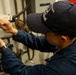 A technician wearing a navy blue uniform and baseball cap is performing maintenance work on a complex pipe and valve assembly. The individual is using two tools simultaneously — a wrench in one hand and what appears to be a screwdriver or pen-style tool in the other. The setting appears to be an industrial or maritime mechanical space based on the aged piping and equipment visible.