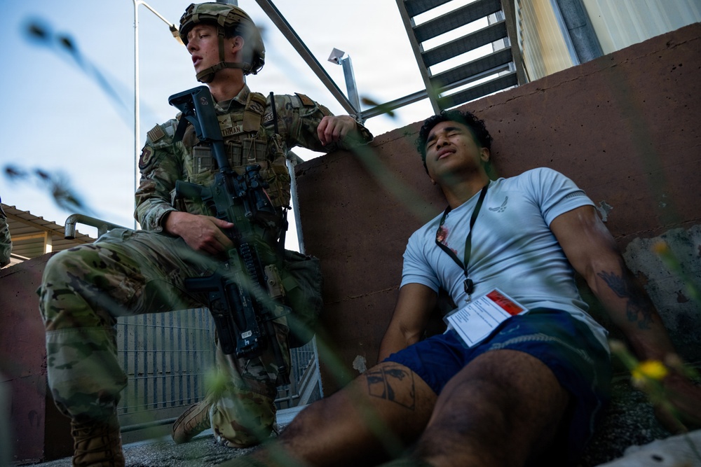 A uniformed military or security personnel in full combat gear, including a helmet and rifle, stands near a civilian who is seated on the ground against a wall. The civilian is wearing a white t-shirt, blue shorts, and has visible simulated injury markings and a lanyard/badge, suggesting this is a training exercise or drill. The setting appears to be an outdoor facility with stairs and a concrete wall.
