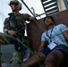 A uniformed military or security personnel in full combat gear, including a helmet and rifle, stands near a civilian who is seated on the ground against a wall. The civilian is wearing a white t-shirt, blue shorts, and has visible simulated injury markings and a lanyard/badge, suggesting this is a training exercise or drill. The setting appears to be an outdoor facility with stairs and a concrete wall.