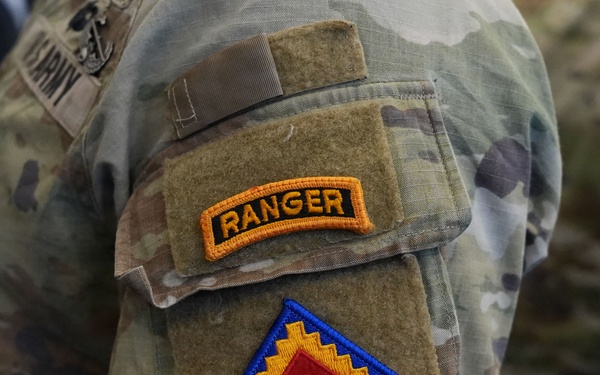 Close-up photograph of a U.S. Army soldier's left sleeve displaying two patches on a MultiCam (OCP) camouflage uniform. The upper patch is a Ranger Tab with gold lettering on a black background, and below it is the U.S. Army Central (ARCENT) shoulder sleeve insignia featuring a blue triangle with gold stepped border and red inner triangle. A 'U.S. ARMY' name tape is partially visible in the background on another soldier's uniform.