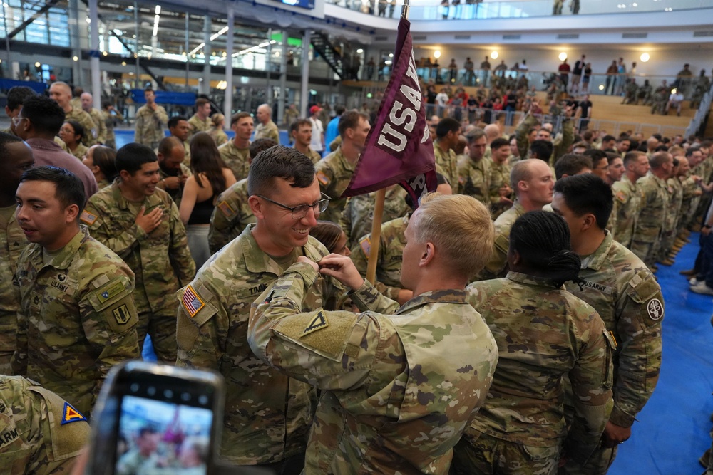 A U.S. Army military ceremony taking place in a large indoor facility, likely a gymnasium or convention center. Soldiers in OCP (Operational Camouflage Pattern) uniforms are gathered in formation while a pinning ceremony appears to be taking place in the foreground. A maroon pennant flag with 'USA' lettering is visible, and observers are watching from an upper-level balcony.