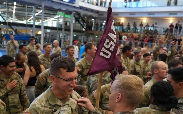 A U.S. Army military ceremony taking place in a large indoor facility, likely a gymnasium or convention center. Soldiers in OCP (Operational Camouflage Pattern) uniforms are gathered in formation while a pinning ceremony appears to be taking place in the foreground. A maroon pennant flag with 'USA' lettering is visible, and observers are watching from an upper-level balcony.