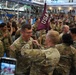A U.S. Army military ceremony taking place in a large indoor facility, likely a gymnasium or convention center. Soldiers in OCP (Operational Camouflage Pattern) uniforms are gathered in formation while a pinning ceremony appears to be taking place in the foreground. A maroon pennant flag with 'USA' lettering is visible, and observers are watching from an upper-level balcony.
