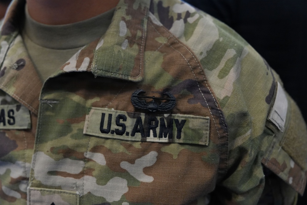 Close-up photograph of a U.S. Army soldier's chest area wearing Operational Camouflage Pattern (OCP) uniform. A 'U.S. ARMY' tape is clearly visible on the right chest, along with a subdued black metal badge above it. A partial name tape ending in 'AS' is visible on the left side of the frame.