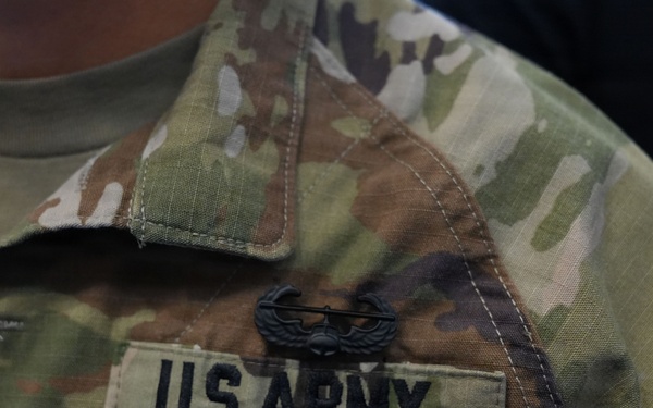Close-up photograph of a U.S. Army soldier's chest area wearing Operational Camouflage Pattern (OCP) uniform. A 'U.S. ARMY' tape is clearly visible on the right chest, along with a subdued black metal badge above it. A partial name tape ending in 'AS' is visible on the left side of the frame.