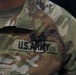 Close-up photograph of a U.S. Army soldier's chest area wearing Operational Camouflage Pattern (OCP) uniform. A 'U.S. ARMY' tape is clearly visible on the right chest, along with a subdued black metal badge above it. A partial name tape ending in 'AS' is visible on the left side of the frame.