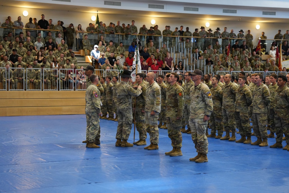 A formal military ceremony is taking place inside an indoor arena or gymnasium with blue matted flooring. Soldiers in MultiCam/OCP camouflage uniforms are standing in formation while a guidon (unit flag) is being presented or transferred. Large crowds of military personnel and civilians fill the bleachers as observers.