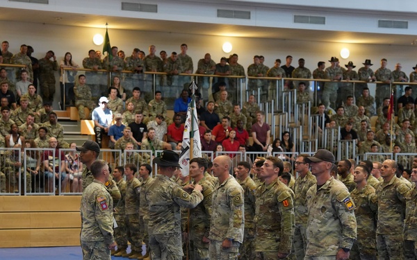 A formal military ceremony is taking place inside an indoor arena or gymnasium with blue matted flooring. Soldiers in MultiCam/OCP camouflage uniforms are standing in formation while a guidon (unit flag) is being presented or transferred. Large crowds of military personnel and civilians fill the bleachers as observers.