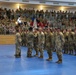 A formal military ceremony is taking place inside an indoor arena or gymnasium with blue matted flooring. Soldiers in MultiCam/OCP camouflage uniforms are standing in formation while a guidon (unit flag) is being presented or transferred. Large crowds of military personnel and civilians fill the bleachers as observers.