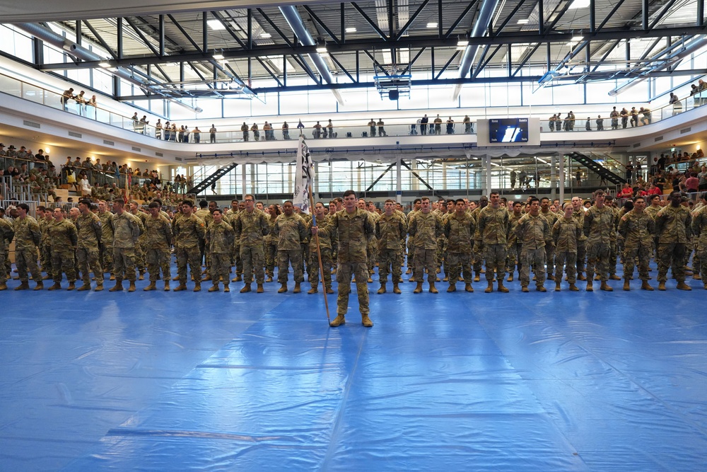 A large formation of military personnel in camouflage uniforms (OCP/MultiCam pattern) stands at attention inside a modern indoor sports facility or gymnasium. A central figure holds a unit guidon flag while rows of soldiers are arranged in precise formation on blue floor mats. Spectators and additional personnel observe from upper balcony levels surrounding the main floor.